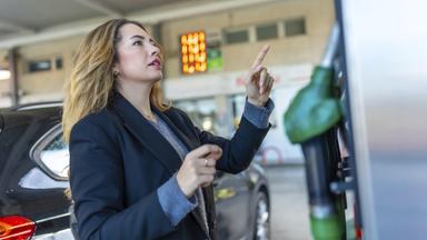 Frau an einer Tankstelle zeigt auf das beleuchtete Preisschild und denkt über steigende Kraftstoffkosten nach, bevor sie ihr Auto betankt, nachdenklicher Pendler, der Budget und Reiseoptionen abwägt