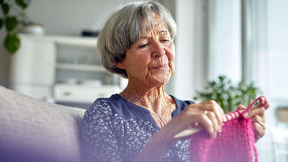 Eine Seniorin sitzt strickend auf dem heimischen Sofa