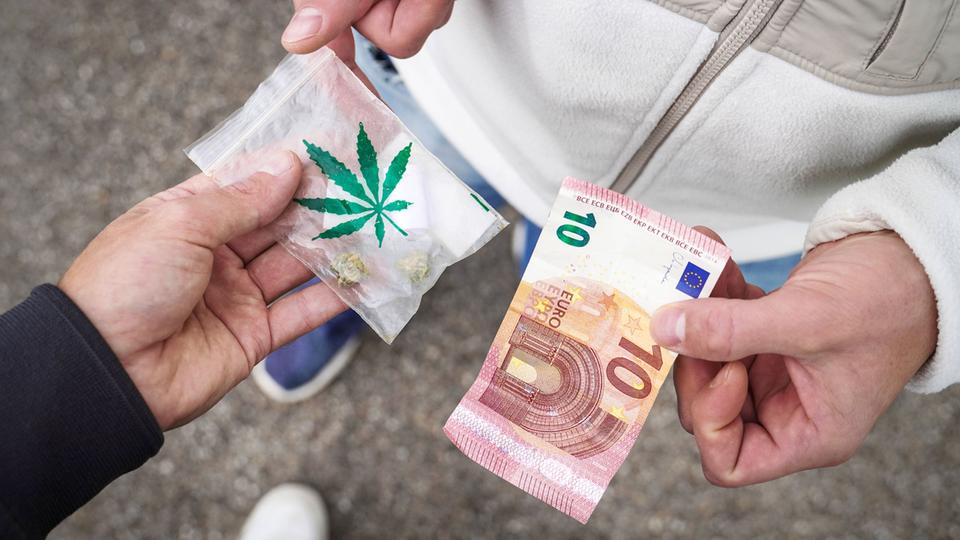 Zwei Männer auf der Straße, einer überreicht eine Tüte Cannabis, der andere überreicht einen 10 Euro Bargeld Geldschein.